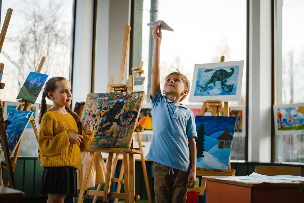 Kids enjoying paper planes amidst colorful paintings in a bright classroom.