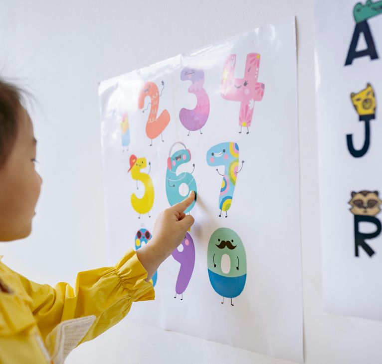 A young girl in a yellow shirt learning numbers in an educational setting, pointing at colorful numbers on a wall poster.
